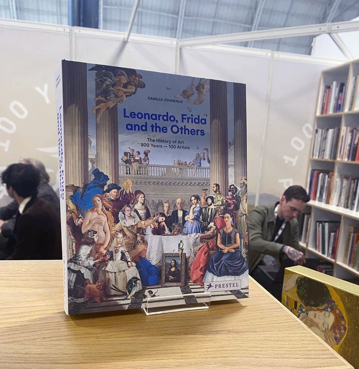 Ein Buch, fotografiert auf der London Book Fair, gestaltet von Weiß Freiburg GmbH für den Prestel Verlag, das Cover zeigt eine Collage verschiedener Evergreens der klassischen und modernen Malerei, unter anderem ist die Venus zu sehen, Frida Kahlo im Selbstportrait, der Schrei von Edvard Munch und die Mona Lisa, diese Figuren sitzen um einen Tisch mit einem antiken Tempel als Hintergrund, der Gesamteindruck ist humoristisch, stilvoll.